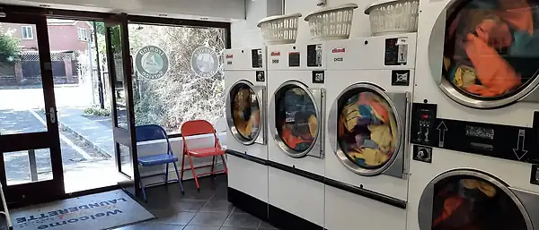 Stacked Cissell dryers tumbling a colourful wash beside the doorway, with red and blue folding chairs and the branded welcome mat.